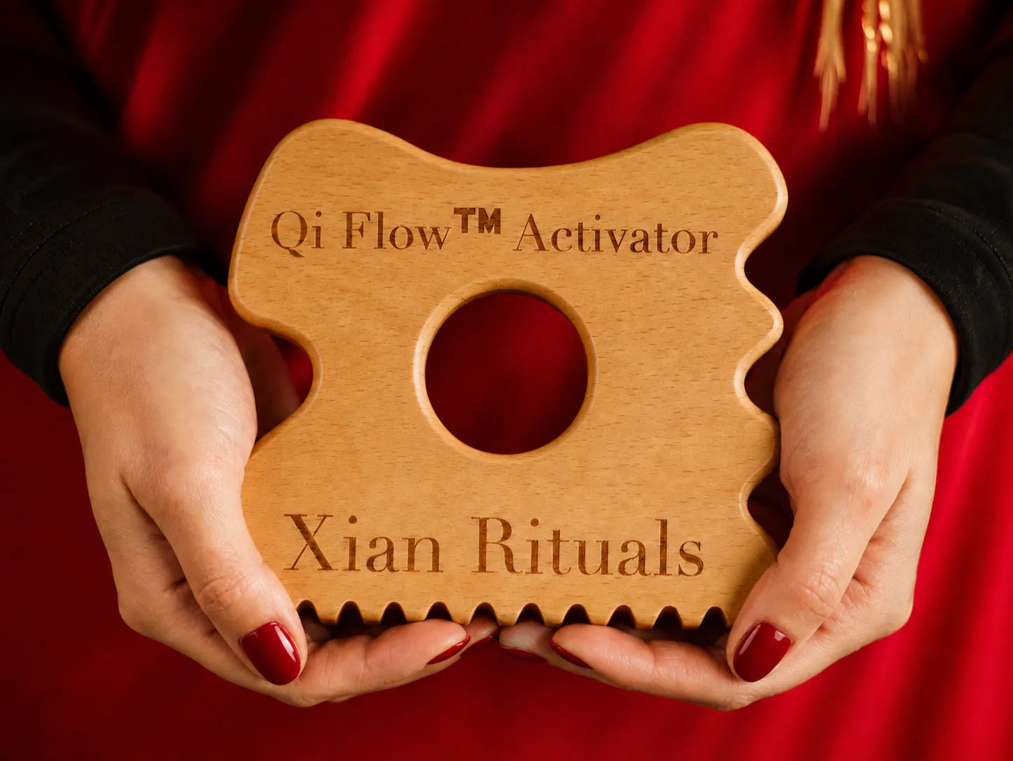 Hands holding Qi Flow Activator wooden gua sha tool against a rich red background